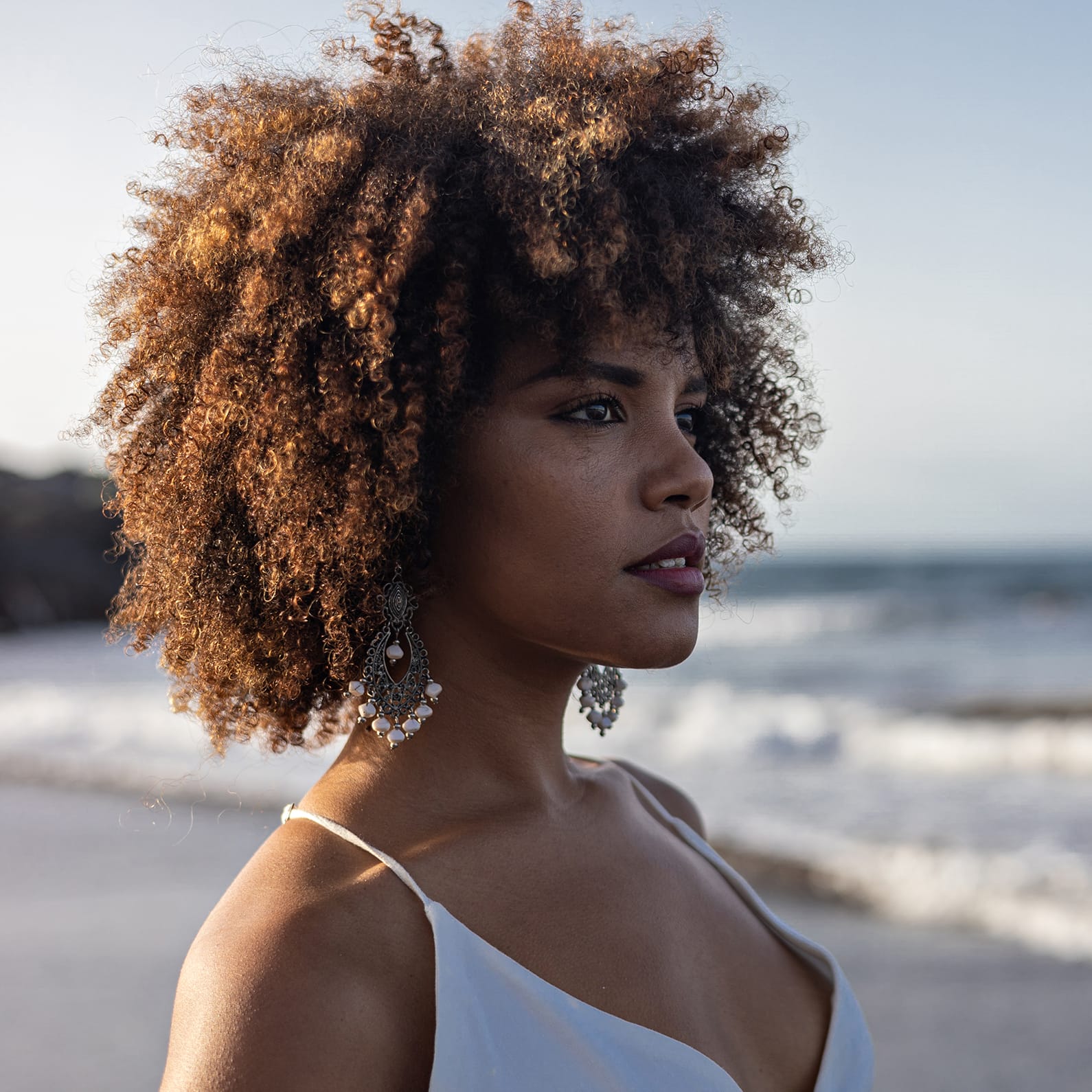 Profile of a woman with curly hair, wearing earrings, by the beach at sunset