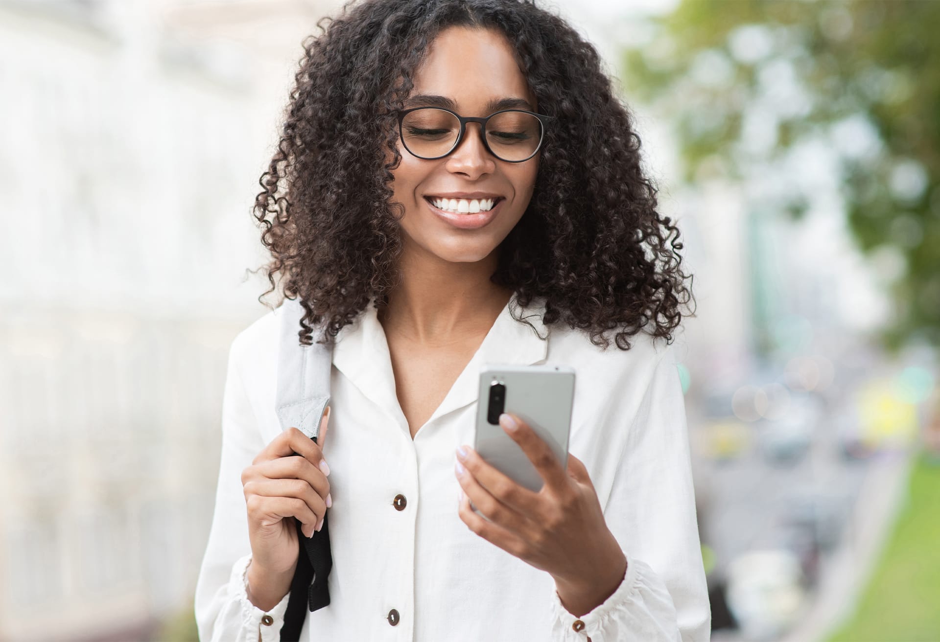 Smiling person with curly hair and glasses holds a smartphone while walking outdoors