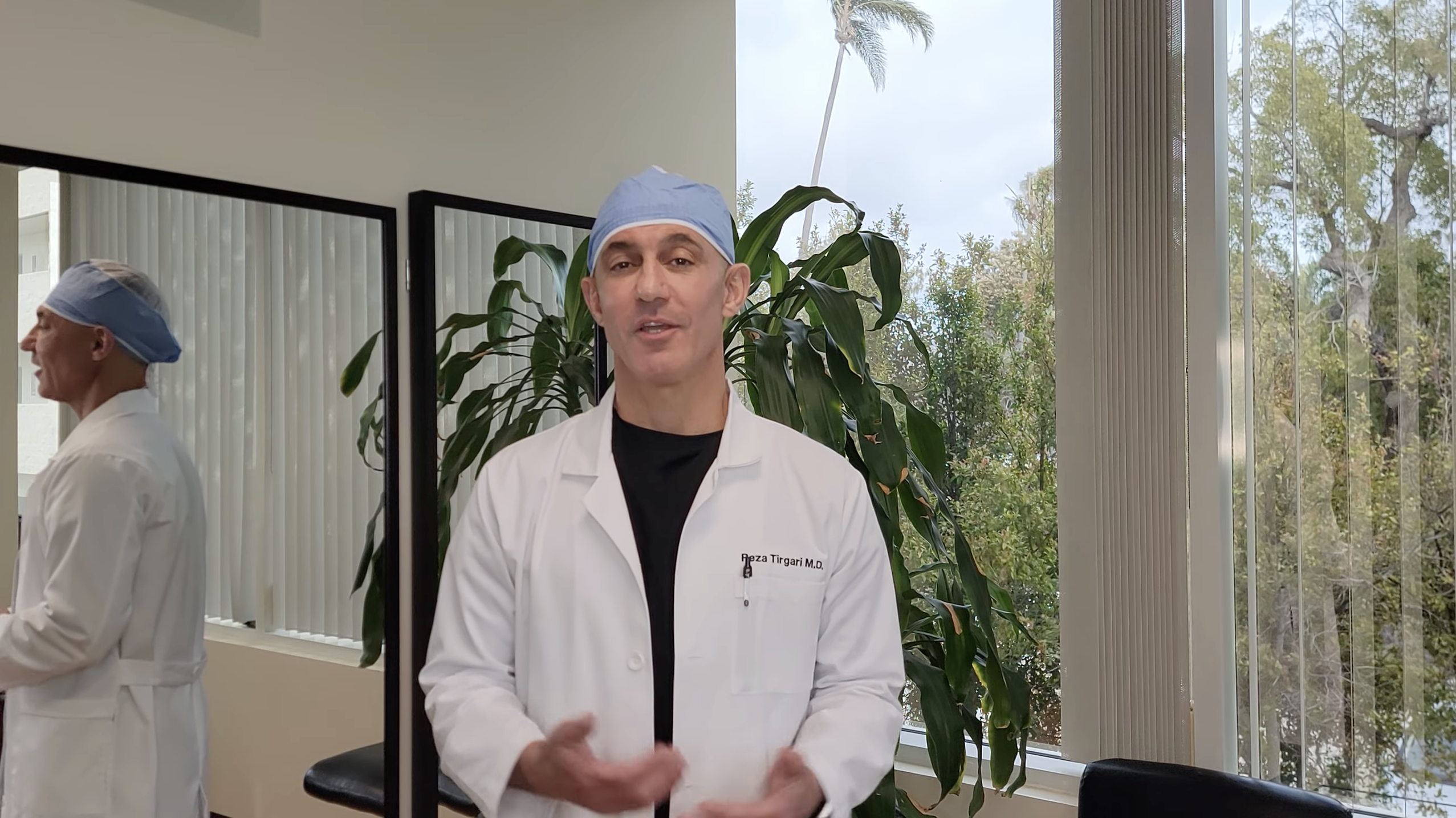 A man in a medical coat and blue cap stands in an office with greenery visible outside