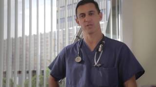 A man in scrubs stands in a well-lit room with vertical blinds. He has a stethoscope around his neck