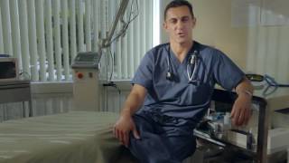 A healthcare professional in scrubs sits on a chair next to a hospital bed