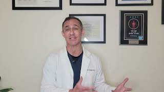 A person in a white coat gestures while speaking in front of framed certificates on a wall