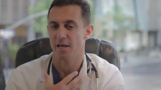 A doctor sits in a chair, discussing something while touching his throat