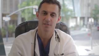 A person in a white coat sits at a desk, looking at the camera with a stethoscope around their neck