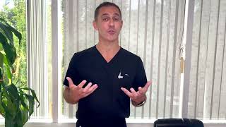 Person in a black shirt speaking with hands raised, standing near a window with blinds