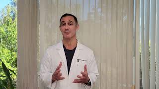 A man in a white coat stands in front of a window, gesturing while speaking