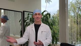 A person in a lab coat gestures while standing in an office with large windows