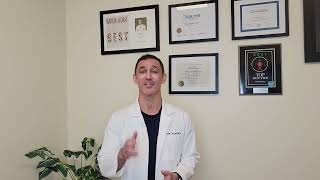 A person in a white coat stands in front of framed certificates on a wall
