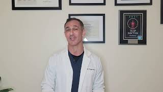 A man in a lab coat stands in front of a wall with framed certificates