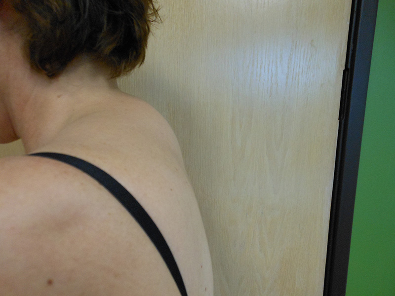 Side view of a person’s shoulder and back with a black strap, near a wall