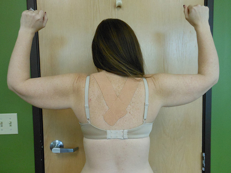 A woman stands with her back to the camera, raising her arms. She wears a beige bra